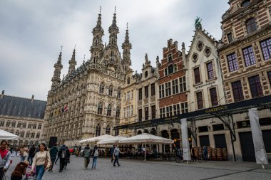 Leuven, Belçika - 12 Ekim 2025: Grote Markt 'taki süslü gotik belediye binasının manzarası, bulutlu bir gökyüzünün altında yürüyen insanlar ve açık hava terasları.