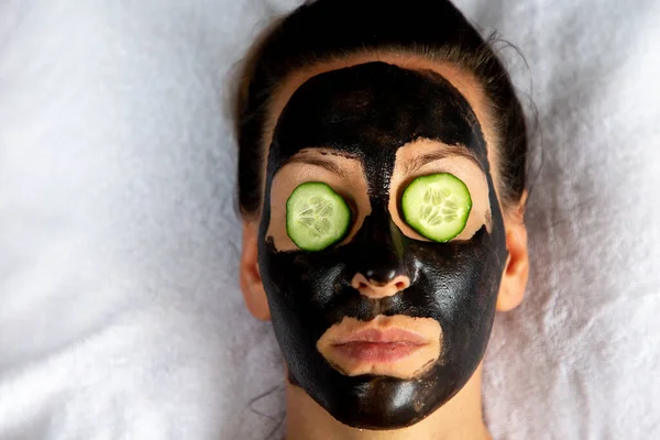 Natuurlijk ogende meisje met zwart caracol gezicht masker leggen op witte handdoek in spa salon Stockafbeelding