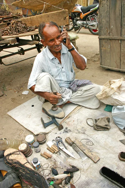 Delhi, Índia - 9 de julho de 2004: Velho indiano conversando em seu celular em sua loja de sapateiros Imagem De Stock