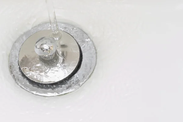 Closeup of tap water flowing into stainless steel drain of white washbasin background — Stock Photo, Image