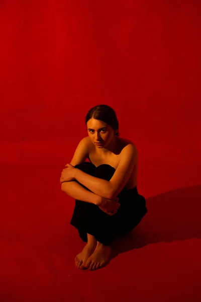 Une fille en pantalon noir étreignait ses genoux, sur un fond rouge — Photo