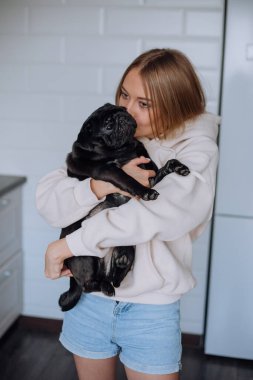 Siyah bir pug ve mavi gözlü sarışın bir kızın portresi, bir köpeği öpüyor.