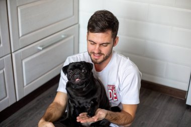 Siyah pug bir adamın yanında oturuyor ve dilini gösteriyor.