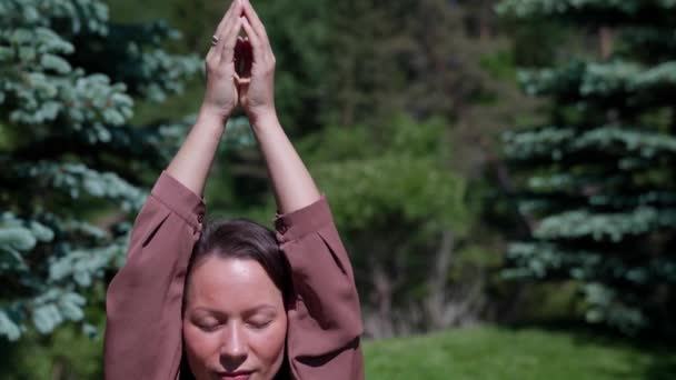 Eine schlanke junge Frau, die in einer Lotusposition sitzt und ihre Hände nach oben in einem Stadtpark hebt. Rückansicht einer Frau, die an einem sonnigen Tag im Freien Yoga praktiziert — Stockvideo