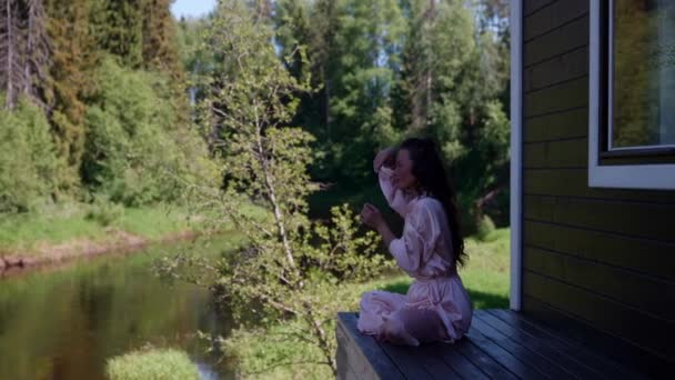 Hermosa joven esbelta sentada en el porche de una casa junto al río tarde soleada en la posición de loto. Un yogui femenino bastante atlético en una asana temprano en la mañana. Aptitud física — Vídeo de stock