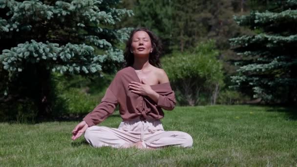 Una mujer practica yoga y medita en la posición de loto en el parque. Chica femenina sana sentada en la posición de loto de sukhasna, meditando o practicando yoga en la naturaleza, al aire libre, relajándose — Vídeo de stock