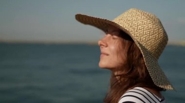 Kafasında büyük bir şapkayla deniz kenarında yürüyen orta yaşlı bir kadının portresi.