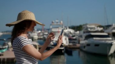Limanda büyük bir şapkayla yürüyen orta yaşlı bir kadının portresi sosyal ağların telefon ekranına bakıyor. Kablosuz teknoloji kullanıcısının modern kavramı.