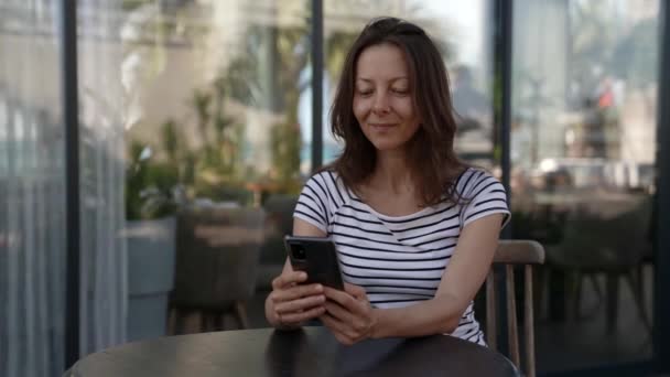 Restoranda bir masada oturan orta yaşlı bir kadının portresi telefon ekranına mesaj yazıyor. Kablosuz teknoloji kullanıcısının modern kavramı. — Stok video
