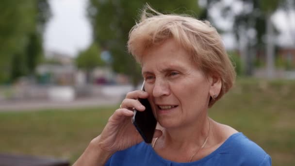 Una signora anziana che usa uno smartphone per parlare, seduta per strada, una donna di mezza età, una nonna che tiene in mano un telefono, godendo della comunicazione in un'applicazione mobile — Video Stock