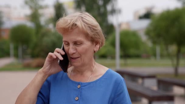 Una donna anziana, che si riposa su una passeggiata, parla su uno smartphone. Una signora anziana eccitata sta chattando per condividere le notizie sul suo cellulare. — Video Stock