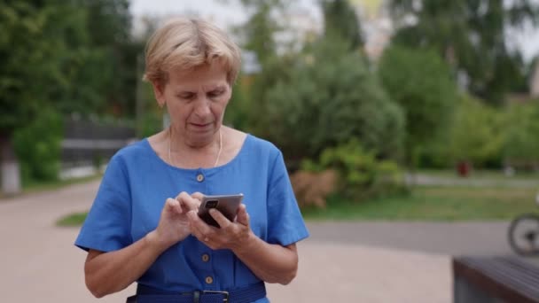 Una nonna anziana si diverte a usare un'applicazione mobile mentre cammina nel parco, digita un messaggio di testo, tiene in mano uno smartphone, guarda lo schermo del telefono cellulare — Video Stock