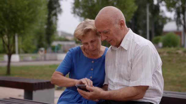 Een bejaard echtpaar geniet van het gebruik van een mobiele applicatie in het park, het vasthouden van een smartphone, kijken naar het mobiele telefoon scherm, browsen op sociale netwerken — Stockvideo