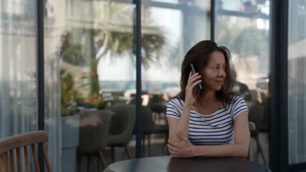 Eine glückliche erwachsene Dame sitzt an einem Tisch auf der Veranda eines Restaurants und telefoniert — Stockvideo