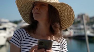 Büyük şapkalı, orta yaşlı güzel bir kadın elinde cep telefonuyla caddede duruyor ve gelen mesajlara cevap veriyor.