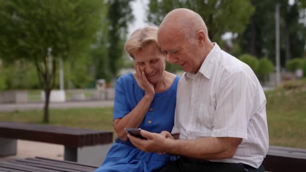 Um casal de idosos apaixonado em um passeio no parque estão assistindo uma mensagem de crianças na tela do telefone móvel — Vídeo de Stock