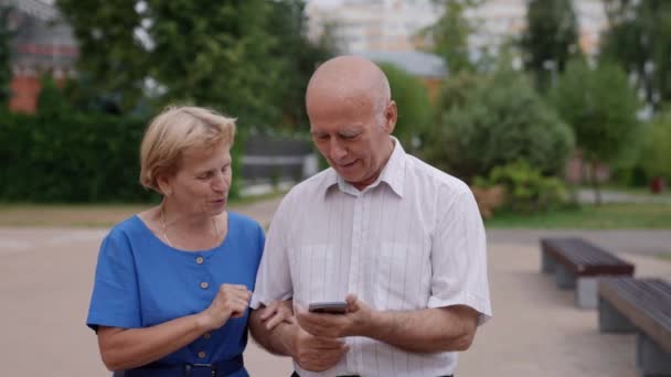 Пожилая супружеская пара, влюбленная во время прогулки, обсуждают новости, которые читают в Интернете, используя мобильный телефон — стоковое видео