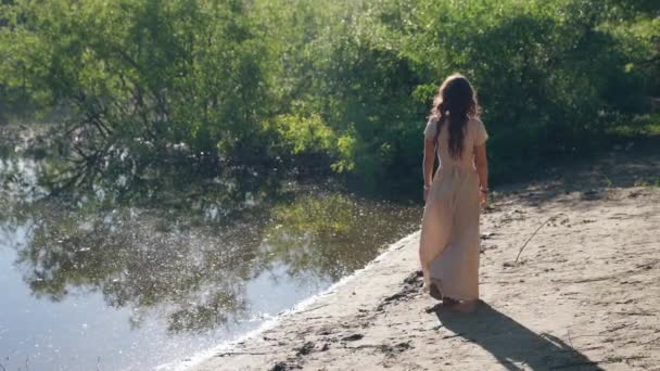 Slow motion video van een jonge vrouw wandelen langs de rivier op het strand in de ochtend in een jurk, een vrouw op vakantie — Stockvideo