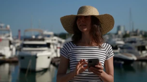 Взрослая женщина в большой шляпе стоит с мобильным телефоном в руке на прогулке к порту яхты — стоковое видео