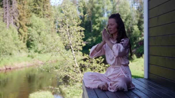 An Asian female yogi is sitting on the veranda with her eyes closed and meditating in the lotus position. Yoga asana for meditation. Early morning. Portrait of a beautiful happy woman. — Stock Video