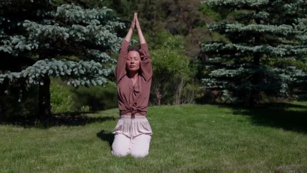 Una yogui asiática se sienta sobre la hierba con los ojos cerrados y practica la respiración. Temprano. Retrato de una hermosa mujer feliz. — Vídeos de Stock