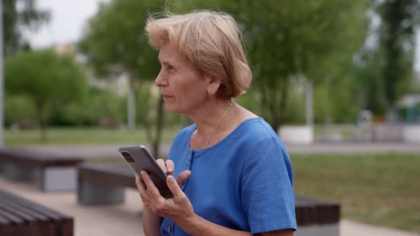 Pojęcie emerytury, komunikacji i osób starszych. starsza kobieta siedzi w letnim parku i pisze wiadomość na swoim smartfonie — Wideo stockowe