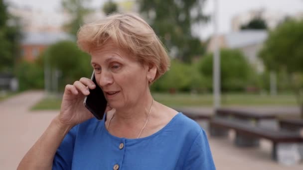 Egy boldog, idős kaukázusi nő sétál a parkban, körülnéz és beszél az okostelefonján. nyugdíjba vonulási életmód kommunikációs technológiákkal otthon — Stock videók