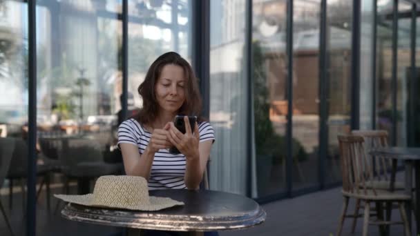 Seorang wanita dewasa yang cantik menggunakan ponsel di kafe yang nyaman. Dia melihat pesan di Internet dan menanggapi pesan teks — Stok Video