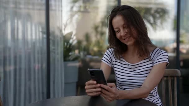 Una mujer adulta envía mensajes de texto, SMS a un teléfono inteligente, se sienta en un café durante el día — Vídeos de Stock