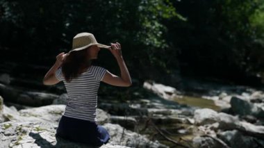 İnce, orta yaşlı bir kadın tatilde, dağlarda dinleniyor. Kafasında şapkasıyla büyük bir taşın üzerinde oturuyor ve temiz hava soluyor.