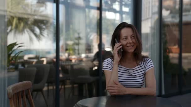 Естественная женщина средних лет разговаривает по телефону в городском кафе, сидящем на веранде — стоковое видео