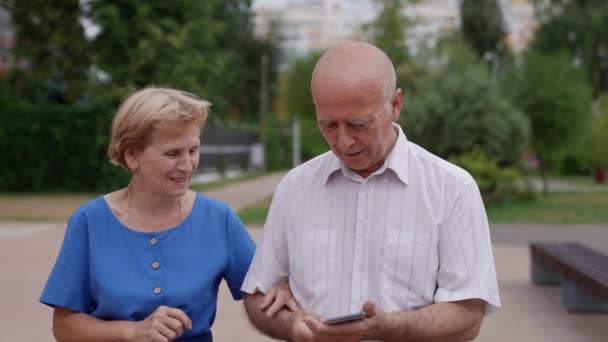 Муж и жена пожилая супружеская пара, гуляющая по улице летом и развлекающаяся просмотром социальных сетей на экране телефона — стоковое видео