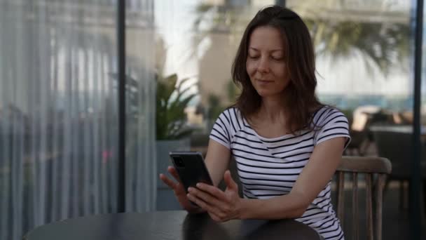 En seriös kvinna sitter på ett café på verandan och väntar på en beställning och tittar på nyheterna på sin telefon — Stockvideo