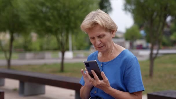 Una anciana con el pelo gris está sentada en un parque con un teléfono móvil viendo mensajes en la pantalla — Vídeos de Stock