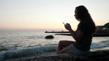 Gün batımında sahilde bir kadının siluetinin yan görüntüsü. Bir kütüğün üzerinde oturuyor ve çevrimiçi iletişim için cep telefonu kullanıyor.
