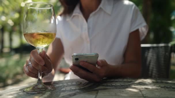 Close-up de mulheres mãos usando o telefone na rua em um café e beber um copo de vinho branco frio — Vídeo de Stock