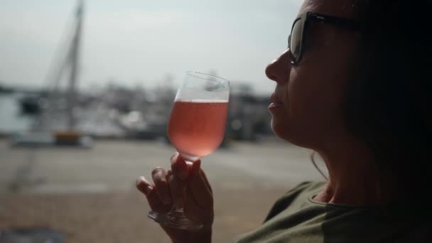 Uma mulher elegante de óculos de sol está sentada na varanda do bar e bebendo uma taça de champanhe rosa — Vídeo de Stock