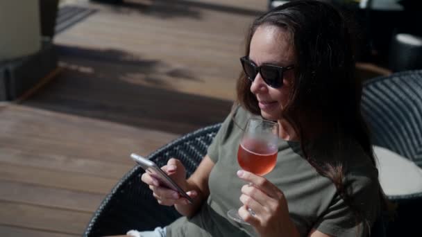 Взрослая привлекательная женщина выпивает коктейль в уличном кафе в солнечный день, используя мобильный телефон для онлайн-общения — стоковое видео