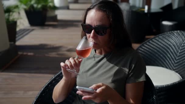 Una mujer hermosa adulta bebe champán rosa en un café de la calle. La chica disfruta del alcohol, relajándose al aire libre. Una señora sentada en una mesa mira la pantalla del teléfono — Vídeo de stock