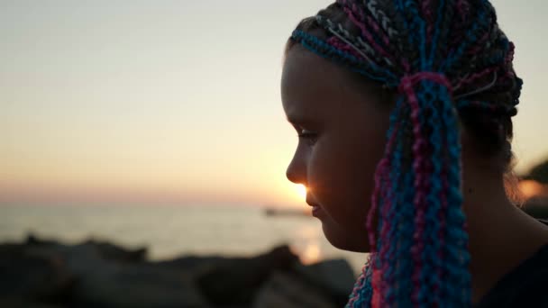 Портретний силует сумної дитини на березі океану в обстановці сонця різнокольорові кіски вплітаються в її волосся — стокове відео