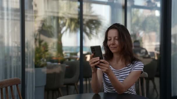 Eine erfolgreiche erwachsene Dame sitzt auf der Veranda eines Cafés und wartet auf das Frühstück und verfolgt die Nachrichten auf ihrem Handy — Stockvideo