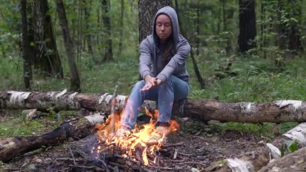 Uma turista adulta está se aquecendo por um fogo aberto sentado em um tronco na floresta — Vídeo de Stock