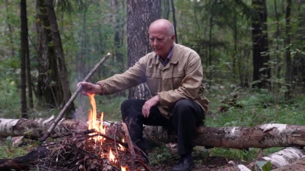 Um velho solitário está descansando na floresta, olhando para um fogo ardente e ajustando a madeira nele, apreciando a natureza e relaxando — Vídeo de Stock