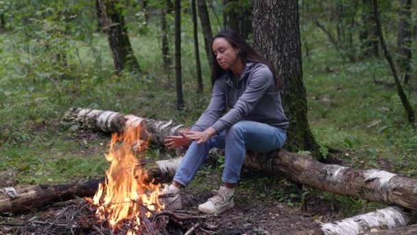 Uma turista adulta está se aquecendo junto ao fogo enquanto está sentada em um tronco na floresta — Vídeo de Stock