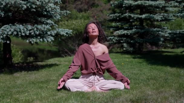 Una mujer practica yoga y respira en la naturaleza sentada en la posición de loto sobre la hierba bajo los rayos del sol diurno — Vídeo de stock