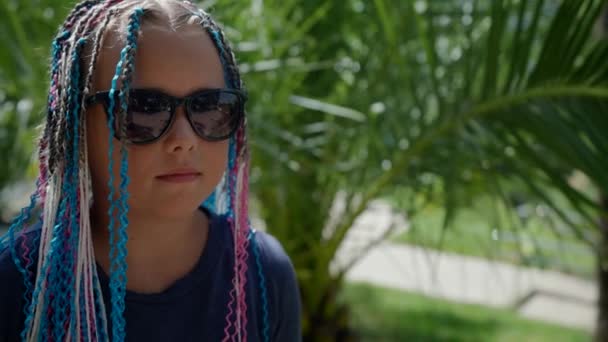 Веселая девочка в солнечных очках на фоне тропического сада с афро-косичками в волосах — стоковое видео