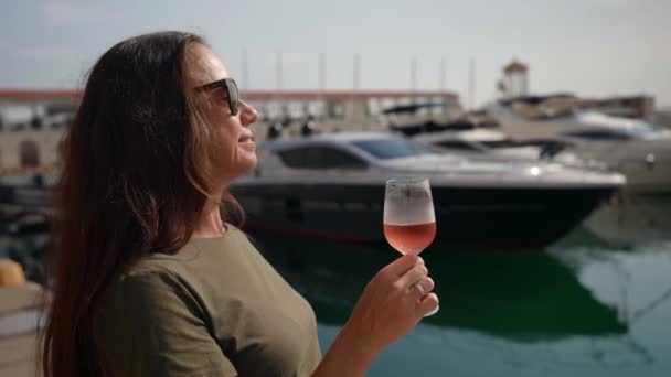 Eine erfolgreiche Frau genießt einen kalten rosa Champagner vor dem Hintergrund des Meeres und einen Hafen mit Yachten — Stockvideo