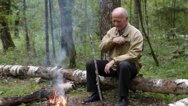 Un turista anciano se está calentando junto a una fogata en el bosque y usa un teléfono celular — Vídeos de Stock