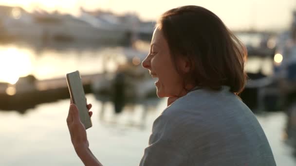 Uma mulher feliz está falando em um link de vídeo para um telefone celular ao ar livre em um fundo embaçado, vista lateral, uma imagem com rastreamento — Vídeo de Stock