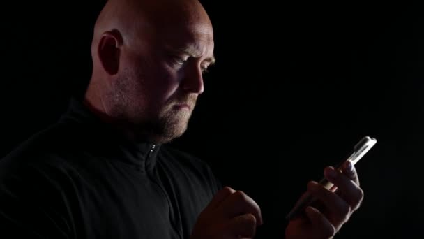 Dospělý holohlavý muž na černém izolovaném pozadí s mobilním telefonem píše textovou zprávu — Stock video